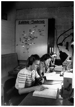 Schwarzweisfoto: Frauen in einem Bunker sitzen an Funkgeräten und schreiben in Bücher. Im Hintergrund ist eine Karte mit leuchtenden Punkten und dem Titel "Luftschutz-Sondernetz" zu sehen. 