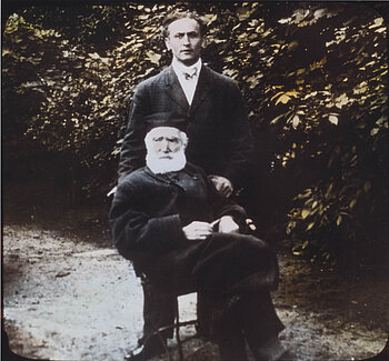 Schwarzweißfoto: Der stehende, junge Harry Houdini steht hinter dem sitzenden, sehr alten Herrn Alexander.