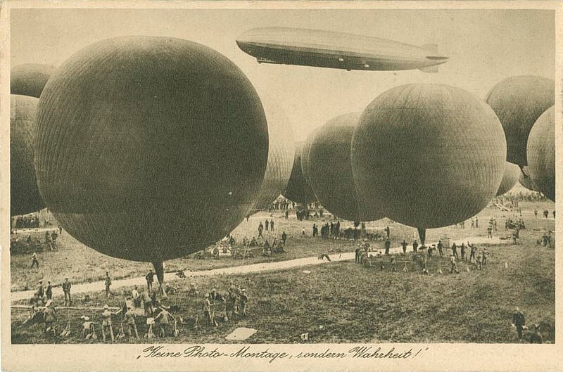 Einige Freiballone am Boden werden von vielen Menschen auf den Start vorbereitet. Im Hintergrund fliegt das Luftschiff Graf Zeppelin.