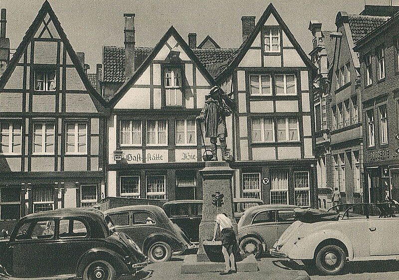 Schwarzweißbild des alten Kiepenkerldenkmals umgeben von Autos, im Hintergrund Fachwerkhäuser.