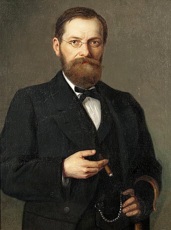 Selbstporträt von Fritz Tüshaus. Ein Mann in mittleren Jahren mit kurzen dunklen Haaren, Vollbart und Brille. Er trägt einen Anzug mit Weste, hält eine Zigarre und einen Schirm.