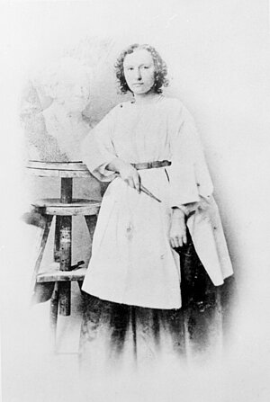 verblassendes Schwarzweißfoto von Elisabet Ney in weißen Kleidern mit kurzen Haaren und mit einem Werkzeug in der Hand. Neben ihr die Büste von Arthur Schopenhauer.