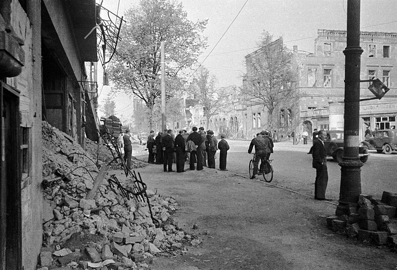 Schwarzweisfoto des Kriegszerstörten Münster. Trümmer auf den Straßen, Menschen, Autos und Fahrräder sind zu sehen. 