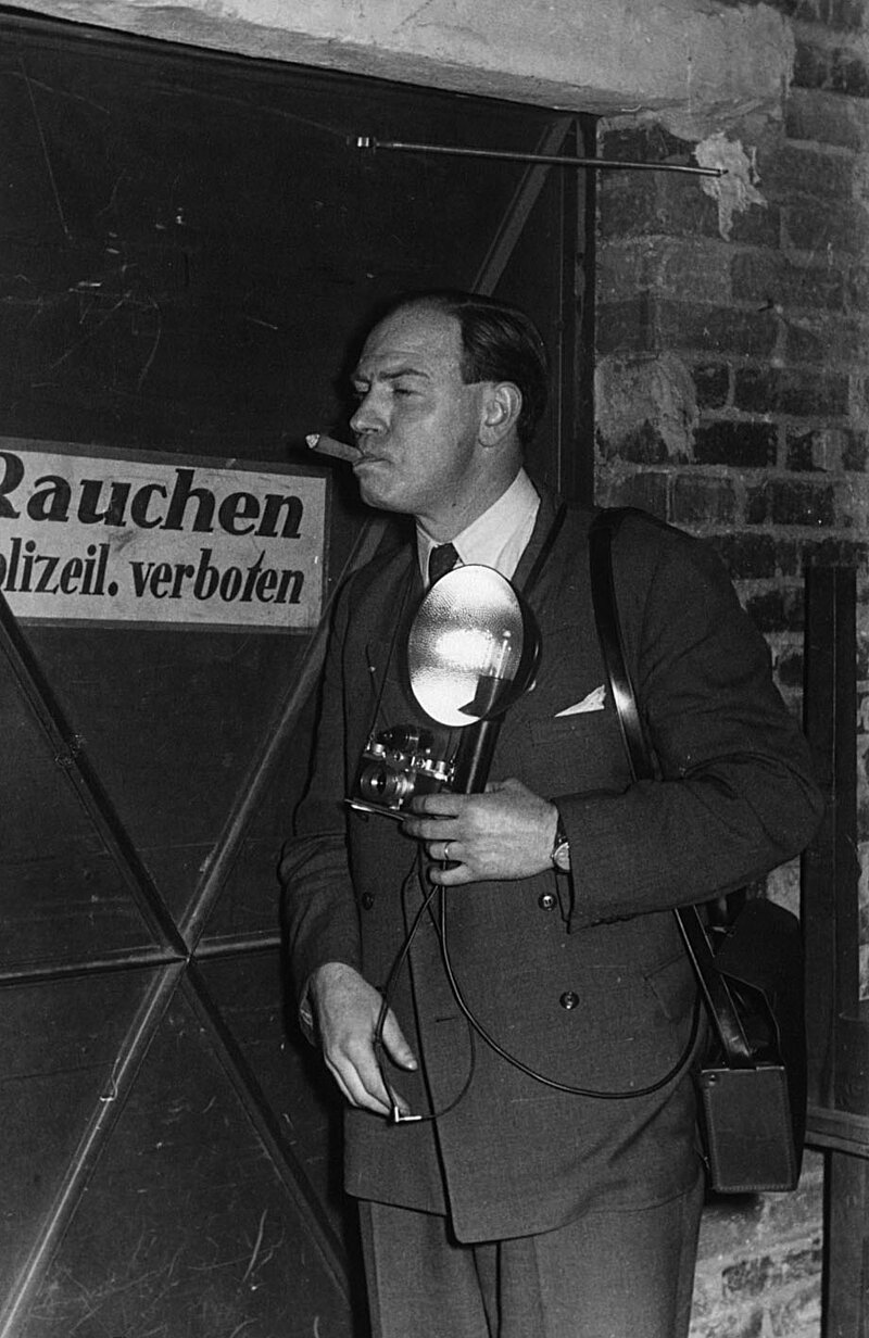 Schwarzweißfoto: Der Fotograf Willi Hänscheid bei der Arbeit. Ein Mann im Angzug mit einer kleinen Kamera mit großem Blitzgerät zieht genüsslich an einer Zigarre. Er steht vor einem Schild auf dem "Rauchen Polizeil. verboten" zu lesen ist.