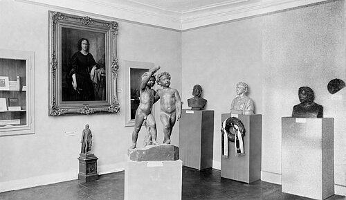 Schwarzweißfoto: Ein Ausstellungsraum mit mehreren Büsten in der rechten Seite, Gemälden auf der linken Seite und einer Skulptur mit zwei nackten Jungen im Zentrum.