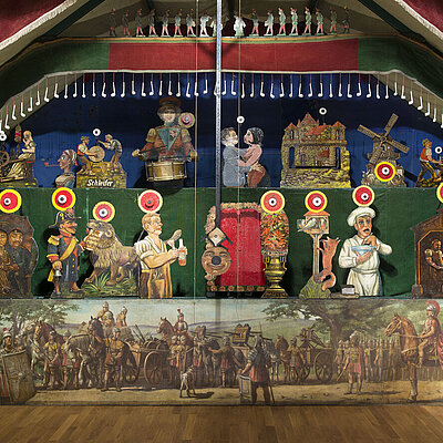 Ein Teil der Schießhalle Schönhagen-Genert. Verschiedene gemalte Figuren von Menschen und Tieren mit Zielen über ihren Köpfen. Darunter ein Banner mit einem Gemälde von römischen Legionären mit Balisten.