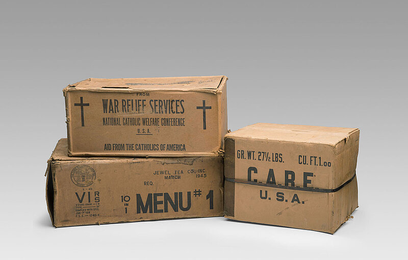 Drei Pappkartons mit den Aufschriften "War Relief Services", "Menu#1" und "C.A.R.E. U.S.A."