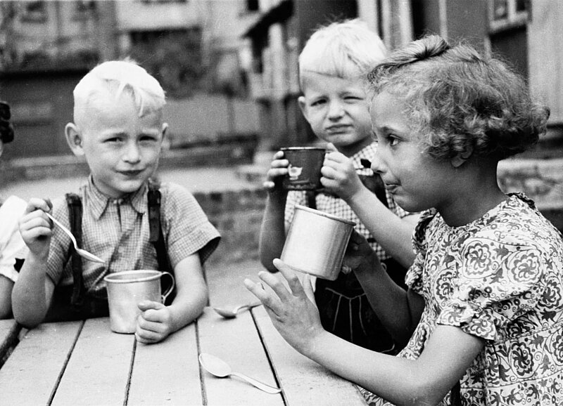 Schwarzweißbild. Drei Kinder essen und trinken mit Löffeln aus Metalltassen.