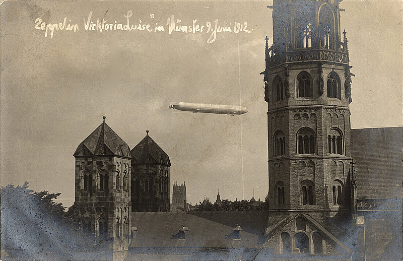 Schwarzweißbild eines Kirchendaches mit drei Türmen. Im Hintergrund ist das Luftschiff Viktoria Luise zu sehen. Text auf dem Bild: Zeppelin VicktoriaLuise" in Münster 9. Juni 1912