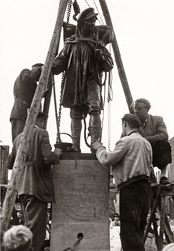 Schwarzweißbild: Vier Arbeiter heben mit einem Holzgestell und Flaschenzügen das Kiepenkerldenkmal auf seinen Sockel. 