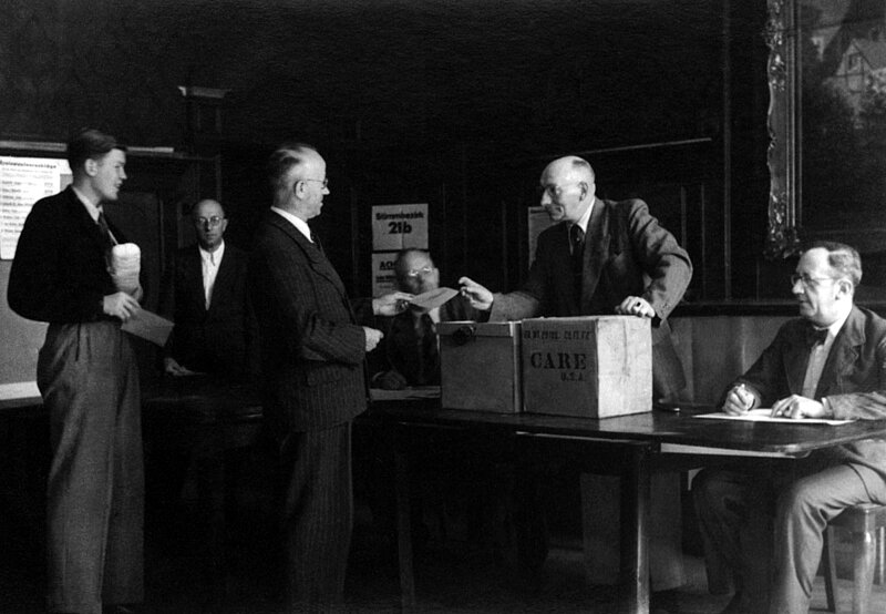 Schwarzweißfoto aus einem Wahllokal, ein Wähler übergibt seinen Stimmzettel an einen Wahlhelfer, der sich auf ein Care Paket stützt, das als Wahlurne dient.