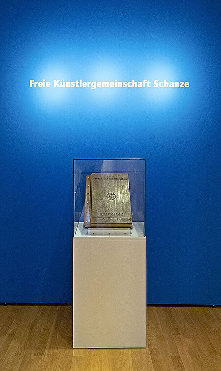 Großes, ledergebundenes braunes Buch in einer Vitrine vor blauem Hintergrund. Darauf der Text "Freie Künstlergemeinschaft Schanze".