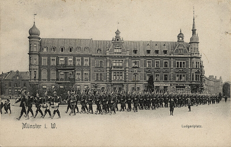 Schwarzweißfoto. Soldaten marschieren in Uniform mit Gewehr und Pickelhaube vor einem Gebäude entlang. 