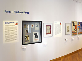 Klenie Plakate und Zeichnungen von Hans Kraft in einer Ausstellung. Daneben Erklärtexte, aufgrund der Auflösung des Fotos unleserlich.