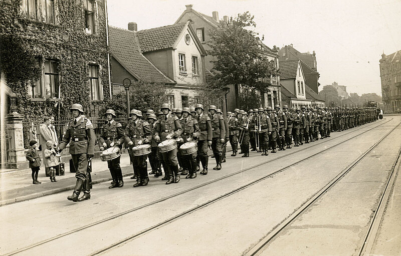 Schwarzweißfoto. Spielmannszug aus Soldaten, dahinter Soldaten mit Waffen in Parade. 